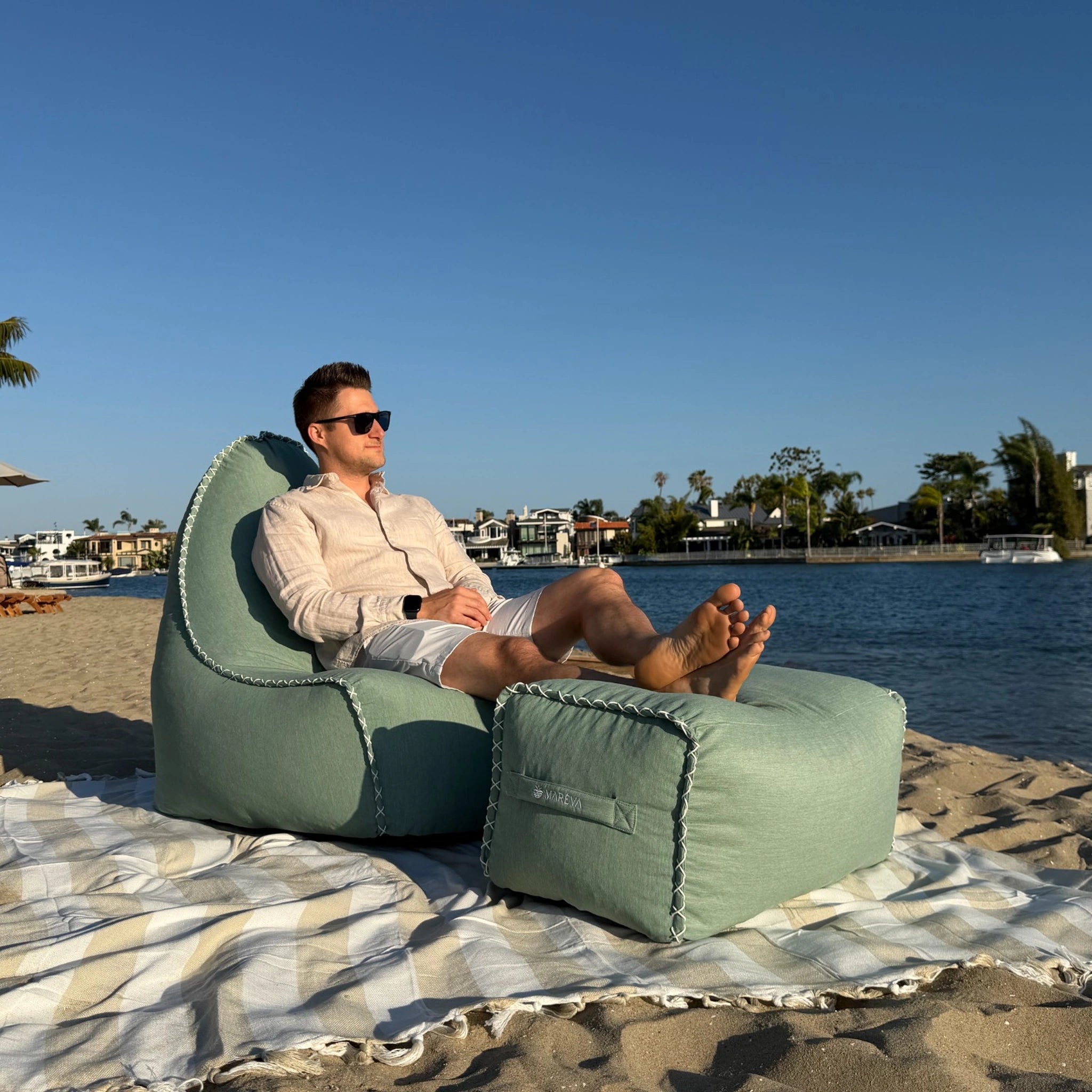 MARÉVA Lounger and footrest pictured in Coastal Sage with male model sitting on it by the harbor.